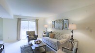This single-floor residence offers a welcoming and well-appointed living area designed with comfort and style in mind. The living room serves as the centerpiece of the space, featuring light-toned walls and smooth ceilings that create an open, airy atmosphere. A large sliding glass door floods the room with abundant natural light while providing direct access to an outdoor area, offering pleasant views of the surrounding greenery.

The living room is thoughtfully arranged around a tufted, neutral-toned sofa accented with decorative throw pillows in earthy and teal hues, complemented by a coordinating wingback armchair. A light-colored rectangular coffee table anchors the seating arrangement atop a patterned area rug, adding texture and warmth to the space. Ambient lighting is provided by classic table lamps positioned on side tables, enhancing the inviting atmosphere during evening hours.

The walls are adorned with framed nature-inspired artwork, adding visual interest and contributing to the calm, cohesive aesthetic of the room. Overall, the space presents a harmonious blend of elegance and everyday comfort, well-suited for relaxed living.