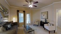 This single-story living space showcases a thoughtfully designed interior that balances comfort and style. The living room serves as the focal point, featuring warm neutral walls accented by elegant crown molding that frames the ceiling. A five-blade ceiling fan with an integrated light fixture anchors the room overhead, providing both function and a classic aesthetic touch.

Natural light enters through multiple windows dressed with horizontal blinds and floor-length draping curtains in earthy tones, complementing the room's warm color palette. A glass-topped coffee table with a chrome frame sits atop plush carpeting, creating a refined centerpiece for the seating area. The furnishings include a dark sofa, a cushioned accent chair, and a chaise, all arranged to encourage conversation and relaxation.

Along one wall, a dark wood console table displays decorative accessories, including a sculptural vase and table lamp, alongside a framed floral artwork that adds a soft, artistic element. A paneled door provides access to an exterior space, further enhancing the room's connectivity to the outdoors. The overall ambiance is warm, calm, and classically styled.