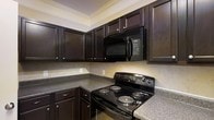 This single-floor space showcases a thoughtfully designed kitchen that serves as a standout feature of the home. The kitchen is outfitted with rich, dark wood cabinetry that extends to the upper walls, providing generous storage while creating a bold, sophisticated aesthetic. The cabinetry is complemented by granite-style countertops in a neutral speckled tone, offering ample workspace for cooking and meal preparation. A suite of black appliances — including an electric coil range and an over-the-range microwave — ties the design together with a cohesive, modern look. The neutral tile backsplash adds texture and depth to the space without overwhelming the dark tones of the cabinetry. Crown molding along the ceiling adds an elegant finishing touch, elevating the overall character of the kitchen. The layout is designed for functionality, with an L-shaped counter configuration that maximizes workspace and ease of movement. The warm, understated color palette throughout creates an inviting atmosphere that balances practicality with style, making the kitchen both a functional and visually appealing centerpiece of the space.