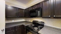 This single-floor space showcases a thoughtfully designed kitchen that serves as a standout feature of the home. The kitchen is outfitted with rich, dark wood cabinetry that extends to the upper walls, providing generous storage while creating a bold, sophisticated aesthetic. The cabinetry is complemented by granite-style countertops in a neutral speckled tone, offering ample workspace for cooking and meal preparation. A suite of black appliances — including an electric coil range and an over-the-range microwave — ties the design together with a cohesive, modern look. The neutral tile backsplash adds texture and depth to the space without overwhelming the dark tones of the cabinetry. Crown molding along the ceiling adds an elegant finishing touch, elevating the overall character of the kitchen. The layout is designed for functionality, with an L-shaped counter configuration that maximizes workspace and ease of movement. The warm, understated color palette throughout creates an inviting atmosphere that balances practicality with style, making the kitchen both a functional and visually appealing centerpiece of the space.