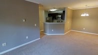 This single-floor residence offers a clean, modern aesthetic defined by an open-concept layout that seamlessly connects the living area to the kitchen. The living space is finished with plush wall-to-wall carpeting and neutral gray walls accented by crisp white crown molding and baseboards, creating a calm and cohesive atmosphere throughout.

The kitchen is a functional centerpiece, elevated by a peninsula with granite countertops that doubles as a breakfast bar and serves as an elegant divider between the cooking and living areas. Dark espresso cabinetry provides ample storage and adds a rich contrast to the lighter tones of the surrounding space. Stainless steel appliances, including a refrigerator and microwave, bring a polished, contemporary finish. A track lighting fixture overhead ensures the kitchen is well-lit and practical.

The open flow between the spaces allows for versatile furniture arrangements and makes the home feel expansive and airy. The consistent use of neutral tones throughout creates a welcoming, move-in-ready environment. Overall, the space strikes a balance between modern design and everyday comfort, making it a practical and stylish place to call home.