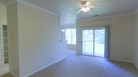This single-floor space offers an open and airy layout highlighted by an abundance of natural light. The main living area features plush carpeting underfoot and is topped with a ceiling fan with integrated lighting, providing both comfort and functionality. Crown molding runs along the upper perimeter of the walls, adding a refined architectural detail to the otherwise clean, neutral interior.

A sliding glass door dominates one wall of the living area, opening to an outdoor balcony that overlooks a lush, green exterior. This feature allows natural light to flood the space throughout the day while offering pleasant outdoor views. A standard window nearby further enhances the brightness of the room.

Transitioning toward the adjacent area, a pass-through counter with a pendant light above it creates a defined yet open connection between the living space and what appears to be a kitchen or dining area. This layout detail promotes an easy flow between spaces while maintaining a sense of distinction between functional zones. The overall ambiance is bright, clean, and inviting, with neutral tones throughout creating a versatile backdrop.