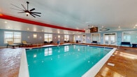 This indoor aquatic facility is thoughtfully designed to provide a comfortable and functional swimming environment year-round. The centerpiece is a generously sized rectangular pool with crystal-clear turquoise water, tiled edges, and entry railings, surrounded by a richly textured stamped concrete deck in warm earth tones. The vaulted ceiling rises impressively overhead, accented by bold red structural beams and large ceiling fans that help maintain air circulation throughout the space. Soft blue walls complement the aquatic atmosphere, while recessed lighting and wall sconces provide consistent, even illumination. Multiple large windows line the perimeter, flooding the interior with natural light and offering views of the surrounding community. Poolside amenities include lounge chairs and seating arrangements positioned along the deck for relaxation. A dedicated lounge area near the entrance features additional seating and large windows. The facility also includes restrooms, a water fountain, and shower access, all conveniently located along one wall. Nautical-themed décor, including a decorative fishing net, oars, and a life preserver, adds a playful maritime character. A double-door entrance with transom windows creates a welcoming entry point, completing this well-appointed indoor pool facility.