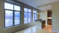 This single-floor urban space showcases a thoughtfully designed open-plan layout that seamlessly integrates the living and kitchen areas. Large, floor-to-ceiling industrial-style windows dominate one wall, flooding the interior with abundant natural light while offering sweeping views of the surrounding cityscape, including neighboring buildings and street-level activity below.

The flooring throughout is light-toned hardwood, lending warmth to the space and complementing the neutral, cream-colored walls. A notable architectural feature is the exposed metallic ductwork that runs along the upper portion of the wall near the windows, adding an industrial loft-like character to the overall aesthetic.

The kitchen area is defined by a substantial island with a dark granite or stone countertop, rich dark wood cabinetry, and an elegant row of pendant lights suspended above on a track rail. The island includes a breakfast bar overhang, providing casual seating. Recessed lighting in the ceiling further enhances illumination throughout the space. The combination of industrial and contemporary design elements creates a sophisticated, urban ambiance well-suited to modern city living.
