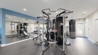 This fitness center is a comprehensively outfitted workout facility designed to support a wide range of training styles and fitness goals. The space is laid out on a single floor and offers a generous open floor plan that accommodates a diverse array of equipment without feeling overcrowded. Durable speckled rubber flooring runs throughout, providing both traction and impact absorption for intense workouts. The walls are painted in a clean combination of neutral gray and accented blue, lending the space a modern, motivating atmosphere. Recessed lighting and ceiling fans ensure the environment remains well-lit and comfortable. A large floor-to-ceiling mirror spans one wall, visually expanding the space and allowing users to monitor their form during exercise. The cardio zone features multiple treadmills, elliptical trainers, and a stationary bike, several of which are equipped with personal display screens. A central multi-functional cable and Smith machine system serves as the strength training hub, complemented by a weight bench and an Everlast heavy punching bag. A water cooler station is conveniently positioned within the facility. Natural light filters in through shuttered windows along one wall, while dedicated doorways provide access to restroom facilities, including a ladies' room. The overall ambiance is clean, professional, and well-maintained.