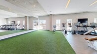 Located at 211 N Fourth Ave in Tucson, Arizona, this fitness center offers a generously sized, single-floor exercise space designed to support a comprehensive range of workout activities. The layout is thoughtfully organized into distinct functional zones, allowing for seamless movement between cardio, strength training, and functional fitness areas.

The facility features a standout turf training zone with bright artificial grass flooring, ideal for agility drills, stretching, and functional movement exercises. Large windows line this area, flooding the space with natural light and offering open views of the surrounding Tucson cityscape. Adjacent to the turf zone, a dedicated cardio section houses a variety of equipment including ellipticals, rowing machines, and stationary bikes, each equipped with personal monitors.

The strength training area is well-appointed with a full selection of selectorized machines, free-weight benches, squat racks, and barbells with plate weights. A dedicated free-weight section features tiered dumbbell racks backed by floor-to-ceiling mirrors, enhancing both the visual depth of the space and functionality for form-checking during workouts.

Architecturally, the space balances industrial and modern aesthetics, with exposed concrete columns and ceilings contrasting against crisp white walls and warm wood-tone flooring. Linear LED lighting runs throughout, providing consistent, even illumination. The overall ambiance is clean, energizing, and purpose-built for serious fitness use.