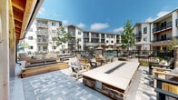 This outdoor kitchen and fire pit area is a thoughtfully designed communal amenity space set within a landscaped courtyard. At its centerpiece stands a substantial rectangular fire table with a stone veneer base and a sleek concrete top, surrounded by Adirondack-style chairs and lounge seating that invite relaxed gathering. The patio surface features a herringbone-patterned stone paver layout that extends throughout the space, lending a polished, cohesive aesthetic underfoot.

Adjacent to the fire pit area, a covered outdoor dining section is framed by exposed heavy timber beams with warm wood-toned ceilings, providing shade and architectural character. This sheltered zone accommodates seating for small groups and is outfitted with wall-mounted lantern lighting for evening ambiance. The building facade features board-and-batten siding with stone base accents, complementing the natural materials used throughout the outdoor space.

Beyond the fire pit area, a wrought iron fence separates the patio from a resort-style pool area, complete with umbrella-shaded seating and pergola structures. The broader courtyard offers lush landscaping with young trees, raised planters, and manicured ground cover, creating a green, inviting atmosphere. Glass double doors connect the outdoor space to interior amenities, ensuring seamless indoor-outdoor flow throughout the property.
