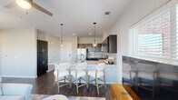 This thoughtfully laid-out single-floor residence offers a comfortable and modern living experience across a well-utilized footprint. The open-concept kitchen and living room serve as the heart of the home, creating a seamless flow between cooking and relaxing spaces. The kitchen is outfitted with dark espresso cabinetry, stainless steel appliances including a refrigerator, range, and microwave, a white subway tile backsplash, and a large island with light granite countertops and bar seating for three. Stylish globe pendant lights hang above the island, adding a contemporary touch. Rich, dark hardwood-style flooring runs throughout the main living areas, grounding the bright, neutral color palette. The living room is generously sized and bathed in natural light from multiple large windows with views to an outdoor balcony, complemented by a ceiling fan. The spacious primary bedroom features plush carpeting, ample natural light, and a sliding barn door that opens to an en-suite bathroom. Two closets provide excellent storage, including a walk-in closet with wire shelving systems. The overall ambiance is clean, modern, and inviting, with an emphasis on functional design and bright, open spaces.
