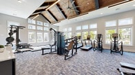The Allora Magnolia fitness center in Magnolia, TX, is a well-appointed, single-floor facility designed to support a comprehensive workout experience. The space is immediately distinguished by its dramatic vaulted ceiling, clad in warm wood planking and supported by exposed dark beams, creating an open and airy atmosphere. A large industrial ceiling fan anchors the ceiling, while linear pendant lighting and recessed fixtures ensure consistent illumination throughout.

Natural light flows generously into the space through a series of large, multi-pane windows lining the perimeter walls, offering views of the surrounding community. The flooring throughout consists of durable, patterned carpet tile in a cool blue-grey palette, providing both comfort and traction during workouts.

The main fitness floor is equipped with a wide range of cardio machines, including treadmills, ellipticals, and a rowing ergometer, arranged to maximize both usability and spacing. A dedicated strength training area features a power rack with a lifting platform, adjustable benches, and an extensive dumbbell rack displayed in front of large floor-to-ceiling mirrors. Additional cable and resistance machines round out the strength offerings.

Entry to the facility is through glass doors branded with the Allora Magnolia name, leading into a light-filled foyer with hardwood flooring and shiplap-accented walls, establishing a polished and welcoming first impression.