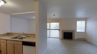 This single-floor residence offers a thoughtfully arranged layout that seamlessly connects the kitchen and living areas. The open living room is carpeted and centered around a clean-faced fireplace, creating a warm and inviting focal point. A large sliding glass door floods the space with natural light and opens to a private balcony, offering scenic views of lush, tree-covered surroundings. The kitchen is positioned just off the main living area and features a double-basin sink, granite-style countertops, and a dishwasher, making it both functional and practical. A structural column partially divides the kitchen from the living space, adding architectural character while maintaining an open feel. The neutral color palette throughout — from the light-toned walls to the beige carpeting — lends the space a calm, versatile ambiance well-suited for a variety of furnishing styles. The combination of natural light, outdoor access, greenery views, and a cozy fireplace makes this a comfortable and well-appointed single-level home.