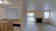 This single-floor residence offers a thoughtfully arranged layout that seamlessly connects the kitchen and living areas. The open living room is carpeted and centered around a clean-faced fireplace, creating a warm and inviting focal point. A large sliding glass door floods the space with natural light and opens to a private balcony, offering scenic views of lush, tree-covered surroundings. The kitchen is positioned just off the main living area and features a double-basin sink, granite-style countertops, and a dishwasher, making it both functional and practical. A structural column partially divides the kitchen from the living space, adding architectural character while maintaining an open feel. The neutral color palette throughout — from the light-toned walls to the beige carpeting — lends the space a calm, versatile ambiance well-suited for a variety of furnishing styles. The combination of natural light, outdoor access, greenery views, and a cozy fireplace makes this a comfortable and well-appointed single-level home.