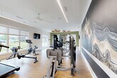 Located at 3616 N Rocky River Road in Monroe, NC, this fitness center offers a generously sized, single-floor exercise room designed to support a full range of workout routines. The space features light hardwood-style flooring throughout, complemented by white walls and accents of sage green that lend a fresh, energizing atmosphere. Linear LED lighting runs along the ceiling, providing bright, even illumination, while a ceiling fan enhances air circulation across the room. Large windows line the perimeter walls, flooding the interior with natural light and offering views of the surrounding outdoor environment. The cardio zone is well-appointed with multiple treadmills, elliptical machines, stair climbers, stationary bikes, and a rowing machine. A dedicated strength training area houses a multi-station cable machine, selectorized weight machines, a power rack with suspension training attachments, and a full rack of dumbbells arranged in front of a large wall mirror with a mounted flat-screen TV. A striking abstract mural in deep navy and grey tones serves as a bold focal point along one wall, adding an artistic dimension to the space. Motivational signage and open floor space for stretching or functional training round out the offering, making this a comprehensive and thoughtfully designed facility.