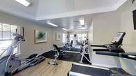 This fitness center is a thoughtfully designed single-floor workout space that balances functionality with a welcoming atmosphere. The room features a combination of hard-surface flooring and rubber matting in the workout zones, providing both comfort and durability for a range of exercises. The layout is spacious enough to accommodate multiple users simultaneously without feeling cramped.

The facility is outfitted with a solid selection of equipment, including treadmills, a rowing machine, adjustable weight benches, and a cable or multi-function strength machine, along with additional cardio equipment visible toward the rear of the space. Equipment is arranged to allow ease of movement throughout the room.

Overhead, flush-mounted fluorescent light panels provide consistent, even illumination throughout the space. Natural light filters in through windows positioned along the far wall, adding brightness and an open feel to the interior. The walls are painted in neutral tones and accented with framed abstract artwork, contributing to a clean and pleasant environment. The overall ambiance is practical yet polished, making it a well-rounded amenity for regular fitness use.