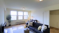 This single-floor space offers a comfortable and well-lit living environment with a thoughtfully arranged layout. The living room serves as the focal point of the space, showcasing classic parquet hardwood flooring that adds warmth and character to the interior. A bank of large, multi-pane windows floods the room with abundant natural light and provides open views of the surrounding urban landscape, creating a bright and airy atmosphere throughout the day. A traditional cast-iron radiator beneath the windows speaks to the classic architectural character of the building. The walls are finished in a clean, neutral white tone, providing a versatile backdrop that complements a variety of décor styles. Flush-mount ceiling fixtures provide consistent overhead lighting. The living area comfortably accommodates a full-sized sofa with accent pillows, a round textured coffee table, and a dark wood console table, demonstrating its generous proportions. The space exudes a relaxed, lived-in charm that makes it both functional and inviting, well-suited for everyday living and casual entertaining alike.
