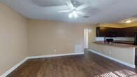 This single-story space is designed with a functional, open-concept layout that connects the living area and kitchen seamlessly. The living room features dark hardwood-style flooring that extends throughout the main living areas, warm beige walls, a textured ceiling, and a ceiling fan with integrated lighting. Large windows allow generous natural light to filter in, brightening the space throughout the day. A floor vent provides climate control, while clean white baseboards offer a crisp contrast to the flooring.

The kitchen is positioned adjacent to the living area and is defined by a peninsula-style countertop with a granite-toned surface, ideal for casual dining or food preparation. Dark espresso cabinetry reaches toward the ceiling, providing ample storage, while black appliances — including a refrigerator, range, microwave, and dishwasher — create a cohesive, modern aesthetic. Hardwood-style flooring continues through the kitchen for a unified look.

The bedrooms are carpeted in a neutral tone and feature warm-colored walls. One bedroom includes a sliding glass door with vertical blinds, offering direct outdoor access and natural light. The bathroom is finished with wood-style flooring, a dark vanity with a granite-topped surface, and a tiled tub and shower combination.