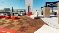 Located at 10130 Foothill Blvd in Rancho Cucamonga, California, this contemporary rooftop terrace offers a well-appointed outdoor retreat with striking views of the surrounding mountains and cityscape under the clear Southern California sky. The space is thoughtfully designed with a mix of materials, combining large-format light-toned tile flooring with warm wood deck tiles that define the primary lounge area. A modern pergola with a translucent roof panel provides partial shade over a comfortable seating arrangement, making it ideal for year-round use. The terrace is furnished with stylish outdoor lounge chairs, modular sofas, and a central fire pit table, creating an inviting social atmosphere. Vibrant red planters serve as bold design accents throughout, filled with drought-tolerant succulents and river rocks, reflecting a Southern California-inspired landscape aesthetic. String lights are strung overhead, adding a warm ambiance for evening gatherings. Concrete and textured panel walls frame the perimeter, contributing to the terrace's modern architectural character, while safety railings ensure open, unobstructed sightlines to the scenic mountain backdrop. A secured utility door and fire safety equipment are also present, underscoring the space's practical design.