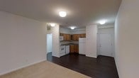 This single-floor residential unit offers a functional and well-organized layout designed to maximize everyday comfort. The open-concept design seamlessly connects the living area and kitchen, creating a sense of spaciousness and ease of movement throughout the space.

The kitchen is fully equipped with warm honey-toned wooden cabinetry, dark stone-style countertops, and white appliances including a refrigerator, range, dishwasher, and over-the-range microwave. The cabinetry provides ample storage, while the layout allows for efficient meal preparation. The kitchen flows directly into the main living area, making it ideal for both daily living and entertaining.

The living area features rich, dark hardwood-style flooring that transitions to plush carpeting, adding warmth and a sense of comfort. Flush-mount ceiling light fixtures provide consistent, even lighting throughout the space. The neutral white walls serve as a clean, versatile backdrop that complements a variety of décor styles.

A hallway visible from the main living space leads to additional rooms, suggesting a well-planned interior layout. The front entry door adds a practical and welcoming point of access. Overall, the unit presents a tidy, move-in-ready environment with classic residential finishes that balance functionality and comfort.