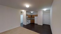 This single-floor residential unit offers a functional and well-organized layout designed to maximize everyday comfort. The open-concept design seamlessly connects the living area and kitchen, creating a sense of spaciousness and ease of movement throughout the space.

The kitchen is fully equipped with warm honey-toned wooden cabinetry, dark stone-style countertops, and white appliances including a refrigerator, range, dishwasher, and over-the-range microwave. The cabinetry provides ample storage, while the layout allows for efficient meal preparation. The kitchen flows directly into the main living area, making it ideal for both daily living and entertaining.

The living area features rich, dark hardwood-style flooring that transitions to plush carpeting, adding warmth and a sense of comfort. Flush-mount ceiling light fixtures provide consistent, even lighting throughout the space. The neutral white walls serve as a clean, versatile backdrop that complements a variety of décor styles.

A hallway visible from the main living space leads to additional rooms, suggesting a well-planned interior layout. The front entry door adds a practical and welcoming point of access. Overall, the unit presents a tidy, move-in-ready environment with classic residential finishes that balance functionality and comfort.
