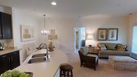 This single-floor residence showcases a thoughtfully designed open-concept floor plan that unifies the kitchen, dining, and living spaces into one cohesive, welcoming environment. The kitchen features a generous curved white countertop with a double stainless steel sink, providing ample workspace and a natural gathering point. Pendant lighting with a classic candelabra-style chandelier illuminates the dining area, where a round table and white chairs create an intimate setting beneath botanical wall art. The living area is carpeted and warmly lit, furnished with wicker and upholstered seating arranged around a central coffee table. A table lamp and framed nature-inspired artwork contribute to the relaxed, coastal ambiance that flows throughout the space. Soft, neutral tones on the walls and ceilings amplify the sense of openness and light. A doorway visible in the background hints at additional rooms, including what appears to be a walk-in closet or bedroom, adding a sense of depth and functionality to the layout. The overall design balances comfort and style with a cohesive, refined aesthetic.