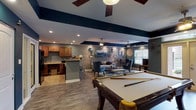The Reserve's community clubhouse is a thoughtfully designed, single-floor amenity space that combines functionality with contemporary style. Throughout the space, rich hardwood-style flooring, bold navy blue accent walls, and recessed lighting create a cohesive and inviting atmosphere complemented by ceiling fans that keep the environment comfortable.

The game area serves as a central focal point, anchored by a full-size billiards table set against the open layout. Adjacent to it, a well-appointed lounge area features a plush sofa, stylish armchairs, and a round wooden coffee table, all framed by an eye-catching gallery wall of circular mirrors and abstract artwork. The seating arrangement is designed to encourage conversation and relaxation.

A fully equipped kitchenette offers warm wood cabinetry, granite countertops, a breakfast bar with modern bar stools, and black appliances, making it well-suited for community gatherings and events. A dedicated workstation area provides a quiet space for residents who need to focus, furnished with a desk and computer setup positioned near natural light from large windows.

The space is accessible through a set of glass-paned double doors that offer views of the surrounding brick exterior. Additional amenities include restroom access and ample storage. Natural light filters in through multiple windows, enhancing the open and welcoming character of this well-rounded community hub.