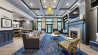 Located at 1 Avalon Dr in Milford, Connecticut, this sophisticated community clubhouse offers a thoughtfully designed single-floor amenity space that blends elegance with functionality. The entrance greets visitors with a striking geometric-patterned tile floor, dark-trimmed French doors, and wainscoting that sets the tone for the refined aesthetic throughout.

The centerpiece lounge area is a showstopper, featuring soaring coffered ceilings, a dramatic gold sculptural pendant light, and a palette of deep navy blues contrasted against crisp white walls. Built-in bench seating flanks a stone-surround fireplace topped with mosaic tile accents, while plush tufted seating and a bold blue area rug create an inviting gathering atmosphere. Large windows allow ample natural light to fill the space.

An adjacent dining and social area offers a communal table and café-style seating, ideal for casual gatherings. A wet bar area with decorative tile backsplash adds an entertainment-ready element to the lounge.

The fitness center is well-equipped with a comprehensive array of cardio machines, cable systems, and strength equipment beneath a coffered ceiling with ceiling fans. A dedicated yoga and group fitness studio features large windows, floor-to-ceiling mirrors, and a full complement of exercise balls, resistance bands, and yoga mats, providing a bright and versatile workout environment.