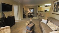 This single-floor residence showcases an open-concept layout that seamlessly connects the living room, dining area, and kitchen into one cohesive living space. The living room is carpeted and generously proportioned, offering ample room for comfortable seating arrangements. A decorative trunk serves as a coffee table centerpiece, adding a rustic charm to the space. Warm beige and tan tones on the walls create an inviting, neutral ambiance throughout.

The dining area sits adjacent to the living room and features a console table with a large mirror, enhancing the sense of depth and light in the space. Bar-height seating with wrought iron stools provides a casual dining option near the kitchen transition.

The kitchen is partially visible from the main living area, elevated slightly with a breakfast bar countertop. Light wood cabinetry lines the upper walls, offering generous storage, while the open pass-through design maintains visual connectivity with the rest of the unit. A pendant light fixture above the dining console adds a decorative focal point. Overall, the space presents a warm, functional, and cohesive interior suitable for everyday living.