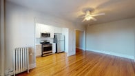 This single-floor unit is thoughtfully laid out, with warm hardwood flooring running continuously throughout every room, lending a cohesive and inviting character to the space. The entry hallway provides a clean transition into the main living areas, with neutral-toned walls and crisp white trim establishing a consistent aesthetic from the moment you step inside.

The open kitchen flows naturally from the living area and is equipped with a full suite of stainless steel appliances, including a gas range, over-the-range microwave, and refrigerator. White shaker-style cabinetry extends to the ceiling, maximizing storage, while light countertops and a single-basin sink complete the functional workspace. A window in the kitchen allows natural light to filter in.

The bedrooms are generously proportioned, each featuring ceiling fans with lighting, neutral walls, and gleaming hardwood floors. Several rooms include windows that bring in natural light and outdoor views. One bedroom features a classic cast-iron radiator alongside a window-unit air conditioner, offering year-round climate control.

The bathroom is finished in a clean, timeless style with white subway tile surrounding the soaking tub and shower, hexagonal mosaic floor tile, a white vanity with a brushed nickel faucet, and a well-lit medicine cabinet mirror above.