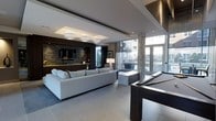 This elegantly designed single-floor space exemplifies contemporary luxury through its open, flowing layout and refined finishes. The main lounge area anchors the space with a generous sectional sofa in crisp white, arranged around a sleek entertainment wall clad in stacked stone tile, complete with a mounted flat-screen television and integrated cabinetry in rich dark wood tones. Recessed ceilings with tiered detailing and designer pendant lighting add architectural depth and a warm, layered ambiance. A full-size billiards table occupies a central position, making a bold statement as a focal point for recreation and entertainment. Floor-to-ceiling windows and glass doors flood the interior with natural light while offering sweeping views of the surrounding outdoor environment, seamlessly connecting the inside to the exterior. Large-format porcelain tile flooring in a neutral tone runs throughout, lending a cohesive and polished aesthetic. A wet bar and additional seating areas provide versatile spaces for gathering and socializing. The overall design balances sophistication with functionality, creating a welcoming environment that is equally suited for relaxed lounging and lively entertainment.