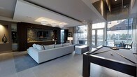 This elegantly designed single-floor space exemplifies contemporary luxury through its open, flowing layout and refined finishes. The main lounge area anchors the space with a generous sectional sofa in crisp white, arranged around a sleek entertainment wall clad in stacked stone tile, complete with a mounted flat-screen television and integrated cabinetry in rich dark wood tones. Recessed ceilings with tiered detailing and designer pendant lighting add architectural depth and a warm, layered ambiance. A full-size billiards table occupies a central position, making a bold statement as a focal point for recreation and entertainment. Floor-to-ceiling windows and glass doors flood the interior with natural light while offering sweeping views of the surrounding outdoor environment, seamlessly connecting the inside to the exterior. Large-format porcelain tile flooring in a neutral tone runs throughout, lending a cohesive and polished aesthetic. A wet bar and additional seating areas provide versatile spaces for gathering and socializing. The overall design balances sophistication with functionality, creating a welcoming environment that is equally suited for relaxed lounging and lively entertainment.