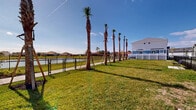 Located at 3784 Bryce Ln in Kissimmee, Florida, this outdoor community space offers a well-maintained, open-air environment designed for leisure and recreation. The grounds feature a striking row of tall, newly planted palm trees lining a paved walking path that runs along the perimeter, adding a distinctly Florida aesthetic to the landscape. Lush, manicured green lawns extend throughout the space, providing ample open area for outdoor activities. A black chain-link fence defines the boundary closest to the water, offering unobstructed views of the adjacent lake, which reflects the surrounding residential neighborhood under the expansive Florida sky. Seating benches are positioned along the lawn, offering comfortable spots to enjoy the waterfront scenery. A white vinyl privacy fence runs along the back boundary, separating the space from the neighboring multi-story residential buildings, which feature a clean, modern exterior in white and blue tones. The overall ambiance is bright and open, benefiting from abundant natural sunlight and wide-open sky views. This thoughtfully landscaped community green space combines functional outdoor amenities with attractive natural surroundings, making it a welcoming area for residents to gather, walk, and relax.
