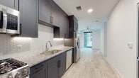 This single-floor unit presents a thoughtfully designed layout with a contemporary aesthetic throughout. The kitchen serves as a standout feature, showcasing dark charcoal shaker-style cabinetry paired with granite countertops and a white hexagonal tile backsplash. Under-cabinet lighting highlights the stainless steel appliance suite, which includes a gas range, built-in microwave, dishwasher, and French door refrigerator. Light-toned hardwood-style flooring runs throughout the main living areas, creating a clean, cohesive look that contrasts with the bold dark cabinetry and interior doors.

A wide, open hallway connects the kitchen to the living area, lined with crisp white walls and recessed lighting overhead. The living space is anchored by a large floor-to-ceiling sliding glass door that floods the room with natural light and provides views of what appears to be a shared courtyard below. A ceiling fan is installed above, adding both function and a finishing detail to the space.

Additional features include a walk-in closet with built-in wire shelving and neutral carpeting, offering practical storage solutions. The overall design balances modern finishes with a clean, minimalist sensibility.