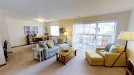 This single-floor residence offers two bedrooms and one bathroom within a thoughtfully arranged floor plan. The open-concept living and dining area serves as the heart of the home, providing generous space for both relaxing and entertaining. The living room features plush carpeting underfoot, a ceiling fan overhead, and a large sliding glass door fitted with vertical blinds that floods the space with natural light and opens to an outdoor area, offering pleasant views and easy access to the exterior. A secondary window further enhances the brightness of the room. The living area comfortably accommodates a full seating arrangement, complemented by a wood-framed coffee table that anchors the space. Adjacent to the living room, the dining area provides room for a full dining table and chairs, making the transition between spaces seamless and practical. A wall-mounted air conditioning unit ensures year-round comfort. The warm, neutral color palette throughout lends the space a welcoming and cohesive ambiance. Overall, the layout is efficient and livable, offering a comfortable setting well-suited for everyday living.