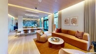Located at 700 Broadway in Santa Monica, CA, this thoughtfully designed amenity space spans a generous single floor, offering a refined blend of indoor comfort and outdoor living. The exterior architecture is defined by floor-to-ceiling glass walls framed in sleek black steel, warm wood cladding, and a clean flat roofline that gives the building a distinctly modern character. Surrounding the structure are multiple landscaped courtyards featuring drought-tolerant succulents, mature olive trees, ornamental grasses, and decomposed granite pathways — creating a serene, California-inspired outdoor environment complete with al fresco seating and outdoor kitchen areas.

Inside, the hallway entry welcomes visitors with large-format travertine tile flooring, a warm wood-paneled ceiling, curated artwork, potted olive trees, and intimate seating nooks. The two living rooms are elegantly appointed, featuring sculptural curved furnishings in soft blue and neutral tones, built-in illuminated shelving, and a cozy banquette — all contributing to a calm, sophisticated ambiance.

The fitness center is a standout space, equipped with a comprehensive array of exercise machines and free weights, set beneath a warm wood-slat ceiling with pendant lighting and expansive windows that open directly to a private outdoor turf area, flooding the room with natural light.