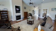 This single-story home offers an open and airy layout designed for comfortable living. The main living area immediately draws attention with its soaring vaulted ceilings, which create an impressive sense of volume and space. A built-in fireplace serves as a focal point along one wall, framed by a clean white mantle and accompanied by a recessed niche housing a flat-screen television — a thoughtful design element that balances function and style. Warm hardwood floors extend throughout the space, lending a cohesive and polished look. Ceiling fans are positioned strategically to maintain airflow across the open floor plan. Natural light filters in generously, brightening the neutral, warm-toned walls that provide a versatile backdrop. A set of classic French doors leads to an adjacent room, offering both visual connectivity and privacy when desired, with additional French doors visible further along — suggesting a series of well-defined yet interconnected living spaces. The living area is anchored by a large area rug with a bold botanical pattern, complementing the casual yet refined furnishings. Overall, the home presents a bright, open, and functional environment with architectural details that add character and charm throughout.