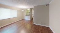 This single-floor unit offers a straightforward and functional layout connecting several key living areas. The main living room is a generously sized space finished with warm hardwood-style flooring and crowned with a textured ceiling. A large window allows natural light to filter in, offering views of surrounding greenery, while a pendant light fixture adds a focal point overhead. Crown molding and clean white baseboards frame the room neatly.

The kitchen is accessible via a pass-through counter with bar-height seating potential, providing an open connection to the living and dining area. It features white cabinetry on both upper and lower levels, a double-basin stainless steel sink with laminate countertops, and space for a refrigerator. The kitchen's layout is compact yet efficient.

The bathroom is well-appointed with white cabinetry vanity, a large mirror framed by bright vanity lighting, wood-look flooring, and a full tub and shower combination. The clean white fixtures create a bright, tidy atmosphere throughout.

The bedroom is carpeted in a neutral tone, providing a comfortable and quiet retreat. A ceiling fan with light kit provides both comfort and illumination, while a standard closet with a hanging rod offers practical storage. A wall-mounted HVAC vent ensures climate control throughout.