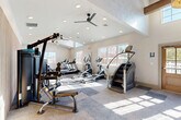 Located at 1070 Lois Ln in Harrisonburg, VA, this fitness center is a thoughtfully designed, single-floor facility offering a spacious and energizing environment for exercise. The space is immediately distinguished by its dramatic vaulted ceilings with exposed wooden beams, which lend a warm, architectural character to the room. A statement chandelier anchors the main entry area, complementing the rustic-modern aesthetic throughout.

Natural light floods the interior through multiple large windows positioned along the walls and a clerestory window above the entrance, creating a bright and welcoming atmosphere. Dual ceiling fans ensure comfortable airflow across the workout floor, which is finished with a patterned, multi-tone carpet tile that adds visual interest and durability underfoot.

The facility is comprehensively equipped for a full-body workout. Cardio offerings include treadmills, ellipticals, stationary bikes, and stair climbers, all positioned to take advantage of the natural light and outdoor views. The strength training area features a multi-station cable machine, adjustable benches, a dumbbell rack, and a large wall mirror that amplifies the sense of space. A vertical slatted wood accent wall adds a refined design touch to the strength zone.

Warm-toned wood double doors with glass panels provide a welcoming entrance, while restroom access and a sanitation station are conveniently available within the space.