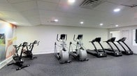 This fitness facility offers a straightforward and functional environment designed to support cardiovascular and general workout needs. The space is contained on a single floor and features a clean, open layout that allows for comfortable movement between equipment. The room is equipped with multiple elliptical trainers and treadmills, providing users with a variety of cardio options. The flooring is covered in dark, rubberized material — a practical choice that provides cushioning and durability for high-traffic use. The ceiling features a suspended tile grid fitted with recessed lighting and ventilation grilles, ensuring the space remains well-lit and properly ventilated during workouts. The walls are finished in a neutral white, contributing to a bright and clean aesthetic. The overall ambiance is utilitarian and purpose-driven, prioritizing function and ease of use. This facility is well-suited for individuals seeking a dedicated space for cardiovascular exercise within a compact, efficiently organized environment.