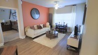This single-floor residence showcases a thoughtfully designed open-concept layout that seamlessly connects the living and dining areas. The living room is anchored by a cream upholstered sofa adorned with earth-toned accent pillows, positioned around a round dark-wood coffee table resting atop a plush ivory shag rug. A bold terracotta-orange accent wall serves as a striking focal point, complemented by a large round decorative mirror that adds visual depth to the space. Wood-style laminate flooring runs throughout, providing a warm and cohesive aesthetic. A ceiling fan with integrated lighting is centrally mounted, contributing both function and style. Natural light pours in generously through large windows dressed with white blinds and sheer curtains, creating a bright and inviting atmosphere. The dining area, situated adjacent to the living space, features a dark wood table paired with matching chairs, making it ideal for everyday meals or intimate gatherings. Decorative branch accents and carefully placed accessories contribute to the warm, contemporary ambiance that defines the overall character of this well-appointed living space.