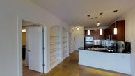 This single-floor space offers a thoughtfully designed open layout that seamlessly connects the living and kitchen areas. A standout feature is the built-in arched shelving unit integrated into the wall, providing both functional storage and a decorative architectural element. The polished concrete flooring runs throughout, lending an industrial yet warm aesthetic to the space.

The kitchen is well-appointed with rich, dark wood cabinetry that contrasts elegantly against stainless steel appliances, including a full-size refrigerator. A kitchen island with a dark stone countertop serves as the centerpiece, accompanied by pendant lighting that hangs at varying heights, adding visual interest and focused illumination. A stainless steel sink is integrated into the island, enhancing its functionality.

Track lighting along the ceiling supplements the pendant fixtures, ensuring the space is well-lit throughout. A corridor visible in the background leads to additional areas of the unit, including what appears to be a doorway with a ceiling-mounted light fixture. The overall ambiance is modern and clean, with a neutral palette punctuated by the warmth of the dark wood cabinetry and polished flooring.