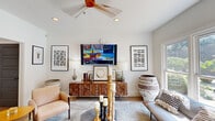 Located in Marietta, Georgia, this single-room living space offers a thoughtfully designed interior that blends contemporary style with eclectic, artistic sensibility. The living room features vaulted ceilings fitted with a striking three-blade ceiling fan and recessed lighting, creating an airy, open atmosphere throughout. Crisp white walls serve as a clean backdrop for a curated selection of bold framed artwork, including geometric and abstract prints that add visual interest to the space. A wall-mounted flat-screen television anchors the media wall above a richly carved wood console, flanked by table lamps and decorative accessories. The seating arrangement includes plush blush accent chairs, a cushioned sofa, and a glass-topped coffee table styled with tall pillar candles. A set of elegant chevron-patterned double wooden doors adds a refined architectural detail to one wall, complemented by gold-accented wall sculptures. Light hardwood flooring runs throughout, grounding the space warmly. A large triple-pane window floods the room with natural light and offers a direct view of an outdoor pool area with lounge seating, seamlessly connecting the interior to the surrounding landscape. A passageway leads to adjacent rooms, hinting at a broader connected layout.