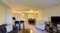 This single-floor residence offers a thoughtfully designed open-concept layout that integrates the main living spaces into one cohesive and functional environment. The kitchen features a breakfast bar with seating for two, pendant lighting overhead, and a pass-through counter that connects it directly to the dining and living areas, making it ideal for entertaining and everyday convenience. The dining area accommodates a round table with multiple chairs and is brightened by a ceiling-mounted light fixture, while colorful wall art adds a welcoming touch. The living area is carpeted and comfortably furnished, providing a relaxed atmosphere anchored by an upholstered accent chair with decorative lettering. The open sightlines throughout the main living space create a sense of roominess and flow, allowing natural light and artificial lighting to reach every corner. Neutral wall tones and warm-toned flooring throughout contribute to a calm, cohesive aesthetic. A hallway visible in the background suggests access to additional private rooms. Overall, the space is well-appointed and functional, balancing open communal areas with a warm, livable ambiance suited for daily life.