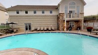 This outdoor recreational space centers around a large, curved swimming pool with clear turquoise water, designed to accommodate a range of aquatic activities. The pool deck is finished with a clean, light-colored concrete surround and is lined with a row of cushioned lounge chairs, providing comfortable spots for relaxation and sunbathing. Safety equipment, including a life ring station, is conveniently positioned poolside.

The adjacent building showcases a distinctive combination of natural stone masonry and light-colored lap siding, creating a warm and welcoming architectural aesthetic. A covered balcony on the upper level overlooks the pool area, while large windows — including prominent upper-floor windows that allow abundant natural light into the interior — contribute to the building's open and airy character. A pitched roof with neutral-toned shingles tops the structure, completing its classic, lodge-inspired design.

Access between the pool area and the building interior is provided through a set of glass-paned double doors, seamlessly connecting indoor and outdoor spaces. The overall ambiance is that of a well-maintained, resort-style facility, blending functional amenities with a comfortable, inviting atmosphere suited to leisure and recreation.