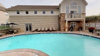 This outdoor recreational space centers around a large, curved swimming pool with clear turquoise water, designed to accommodate a range of aquatic activities. The pool deck is finished with a clean, light-colored concrete surround and is lined with a row of cushioned lounge chairs, providing comfortable spots for relaxation and sunbathing. Safety equipment, including a life ring station, is conveniently positioned poolside.

The adjacent building showcases a distinctive combination of natural stone masonry and light-colored lap siding, creating a warm and welcoming architectural aesthetic. A covered balcony on the upper level overlooks the pool area, while large windows — including prominent upper-floor windows that allow abundant natural light into the interior — contribute to the building's open and airy character. A pitched roof with neutral-toned shingles tops the structure, completing its classic, lodge-inspired design.

Access between the pool area and the building interior is provided through a set of glass-paned double doors, seamlessly connecting indoor and outdoor spaces. The overall ambiance is that of a well-maintained, resort-style facility, blending functional amenities with a comfortable, inviting atmosphere suited to leisure and recreation.