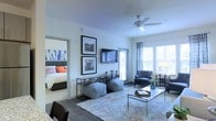 This single-floor residence showcases a thoughtfully designed open-concept layout that seamlessly connects the living area to the adjacent bedroom. The living room serves as the centerpiece of the space, featuring smooth white walls adorned with framed artwork and a wall-mounted flat-screen television positioned above a sleek metal console. A ceiling fan with a light fixture adds both functionality and a clean, contemporary touch overhead.

The flooring throughout the main living area consists of rich hardwood-style planks, anchored by a vibrant, patterned area rug that adds warmth and color to the space. Furnishings include a pair of plush gray poufs, a glass-top coffee table, and a comfortable accent chair, all contributing to a modern, coordinated aesthetic.

Natural light floods the room through large windows dressed with patterned drapes and horizontal blinds, while a glass door provides direct access to an outdoor area, enhancing the sense of openness. The adjacent bedroom, visible through an interior doorway, features plush bedding with bold accent pillows and a tufted upholstered headboard, completing this well-appointed residence.