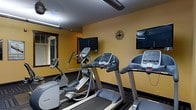 This fitness center is a purpose-built exercise space designed to meet essential cardio and workout needs. The room features warm yellow-toned walls that create an energizing atmosphere, complemented by dark patterned carpeting that adds a structured, functional aesthetic throughout. The space is outfitted with a suite of Precor cardio equipment, including treadmills, an elliptical trainer, and a stationary bike, all arranged to allow comfortable use without overcrowding. Multiple flat-screen televisions are mounted on the walls, allowing users to stay entertained or informed during their workouts. A large wall mirror on one side of the room enhances the sense of space while allowing users to monitor their form. A decorative wall clock adds a practical touch, helping users track their workout duration. Overhead fluorescent lighting ensures the room is well-lit throughout. A window provides a visual connection to the outside, contributing to a less confined feel. The overall design is straightforward and utilitarian, prioritizing function and ease of use, making it a convenient option for those seeking a reliable space for daily cardio exercise.