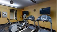 This fitness center is a purpose-built exercise space designed to meet essential cardio and workout needs. The room features warm yellow-toned walls that create an energizing atmosphere, complemented by dark patterned carpeting that adds a structured, functional aesthetic throughout. The space is outfitted with a suite of Precor cardio equipment, including treadmills, an elliptical trainer, and a stationary bike, all arranged to allow comfortable use without overcrowding. Multiple flat-screen televisions are mounted on the walls, allowing users to stay entertained or informed during their workouts. A large wall mirror on one side of the room enhances the sense of space while allowing users to monitor their form. A decorative wall clock adds a practical touch, helping users track their workout duration. Overhead fluorescent lighting ensures the room is well-lit throughout. A window provides a visual connection to the outside, contributing to a less confined feel. The overall design is straightforward and utilitarian, prioritizing function and ease of use, making it a convenient option for those seeking a reliable space for daily cardio exercise.