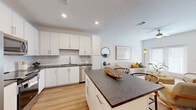 This single-floor residence is designed with a clean, contemporary aesthetic and a well-considered open-concept layout. The kitchen is a standout feature, outfitted with crisp white cabinetry extending to the ceiling, dark granite-style countertops, and a full suite of stainless steel appliances including a refrigerator, range, microwave, and dishwasher. A central island with seating provides additional prep space and a natural gathering point, seamlessly connecting the kitchen to the living area. Light wood-style flooring runs consistently throughout, enhancing the sense of continuity and warmth.

The living area is bright and airy, benefiting from natural light through a set of glass French doors that open outward. A ceiling fan overhead adds to the comfort of the space. The layout accommodates a comfortable seating arrangement and media area, offering a relaxed yet functional environment.

The bedroom features a bold green accent wall that adds depth and character, complemented by a large window with blinds that allows for ample natural light. A ceiling fan is also present in this room.

The bathroom maintains the same design language as the rest of the residence, with white cabinetry, dark countertops, an undermount sink, and wood-style flooring, creating a cohesive and polished finish throughout the home.