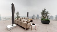 This sophisticated rooftop terrace occupies the uppermost level of a high-rise building, offering sweeping panoramic views of a dense urban skyline. The terrace is finished with large-format porcelain tiles that lend a clean, contemporary aesthetic across its generously proportioned open-air footprint. A glass-panel perimeter railing preserves unobstructed sightlines to the cityscape below, allowing natural light to permeate the space throughout the day.

The main lounge area is anchored by a plush, neutral-toned sectional sofa paired with sleek marble-top accent tables and a central rectangular fire pit, creating an inviting setting for gatherings. Wire-frame Acapulco-style chairs and side tables provide additional flexible seating arrangements. A row of contemporary white chaise lounges lines one side of the terrace, offering a dedicated relaxation zone.

Mature olive trees and lush plantings in large gray concrete planters are strategically positioned to add privacy and a natural contrast to the building's modern metal-panel façade. Decorative succulent planters add additional texture and color throughout. Utility access points, including a window washing equipment room and exit doors, are neatly integrated into the rooftop structure without disrupting the terrace's refined atmosphere.