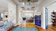 Located in Raleigh, NC, this thoughtfully designed single-floor community clubhouse offers a generous, open layout filled with upscale finishes and a cohesive blue-and-gold color palette throughout. The entry foyer welcomes visitors with warm hardwood flooring, a striking geometric-patterned area rug, and a bold sculptural chandelier featuring cobalt blue and gold accents beneath a coffered ceiling detail. The expansive living room beyond serves as the heart of the space, highlighted by soaring double-height ceilings, floor-to-ceiling sliding glass doors, and abundant natural light that pours in from oversized windows framing views of the outdoor courtyard and pool area. A sleek linear fireplace anchors one wall, flanked by built-in shelving and curated blue-toned artwork, while plush tufted blue velvet armchairs and a curved loveseat create inviting seating arrangements. A separate lounge area features a bold geometric-patterned carpet, a glass-top dining table with gold-accented chairs, and additional seating nooks ideal for meetings or casual gatherings. The clubhouse also includes a well-appointed coffee and refreshment bar with stone tile flooring, white cabinetry, a kitchen island with bar seating, and direct access to the outdoors. A dedicated fitness room provides spin bikes, smart parcel lockers, and natural light from large windows, rounding out this vibrant and functional community hub.