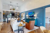 This thoughtfully designed single-story space functions as a welcoming community or leasing hub, blending coastal-inspired aesthetics with modern functionality. Throughout the interior, herringbone-patterned hardwood flooring creates a striking visual foundation, while soaring vaulted ceilings with exposed dark wood beams lend an airy, grand quality to the main areas.

The reception area greets visitors with shiplap-paneled walls, a built-in front desk, and an abundance of natural light. Adjacent private offices offer a quieter, more focused setting for consultations. A bold blue shiplap accent wall defines the central lounge, which features white upholstered seating, leather accent chairs, a mounted flat-screen television, and statement pendant lighting.

The wet bar area is appointed with slate-blue subway tile, open floating shelves, white shaker cabinetry, dark countertops, a beverage cooler, and a built-in bar with tufted bar stools. A sunlit dining nook with floor-to-ceiling sliding glass doors and garden views provides a casual gathering space. Decorative shiplap walls, curated greenery, and a neon logo sign complete the polished, resort-inspired ambiance throughout.